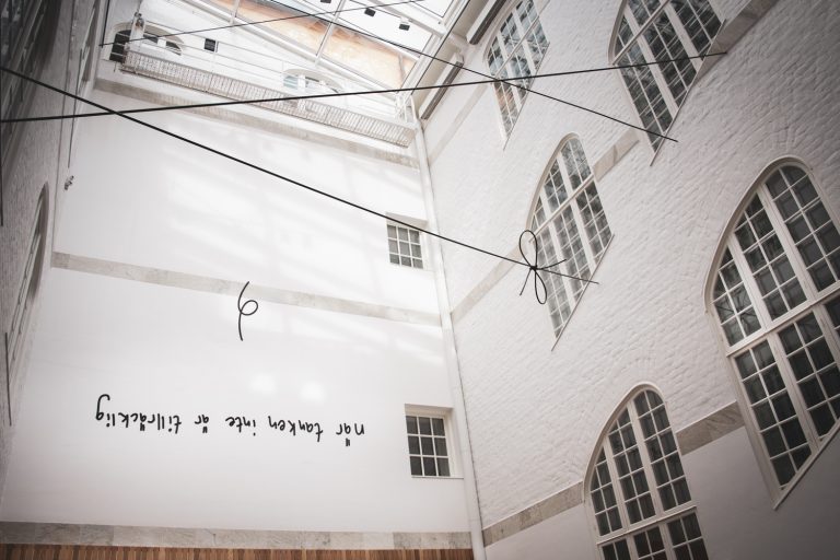 En innergård med glastak. På väggen finns bokstäver och svarta linjer syns ii luften genom rummet.