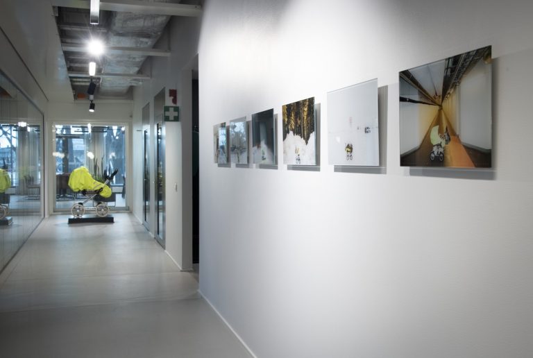 En gul barnvagn i bakgrunden och sex stycken fotografier som hänger på en vägg.